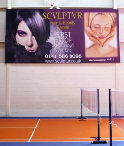 Photo shows a full-colour advertising Billboard Poster for Sculptur Hair & Beauty Salons in an indoors tennis court at David Lloyd health club in Glasgow. The poster has two portraits of models, one for hair and one for beauty treatments.
