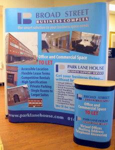Photo shows a curved wall-type of exhibition stand for Broad Street Business Complex in Glasgow. It has a sky blue (cyan) background with exterior and interior photos of the business centre, committee room, foyer etc. In front of the curved wall is the transit case it travels in, dressed with a matching graphic wrapper and a wooden counter-top to make it a counter during the exhibition.
