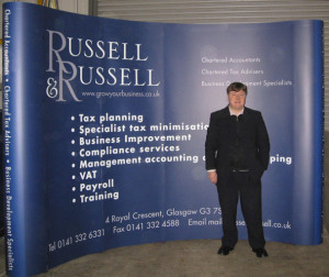 Picture shows a four-by-three size exhibition stand I designed for Russell & Russell Accountants. Wider than most exhibition stands, it's a dark blue curved wall with the R&R logo top left, white bulletpoint text in the centre and contact information along the foot. The curved endcap corners have a lighter shade of blue. I'm shown standing in front of the stand in the workshop.