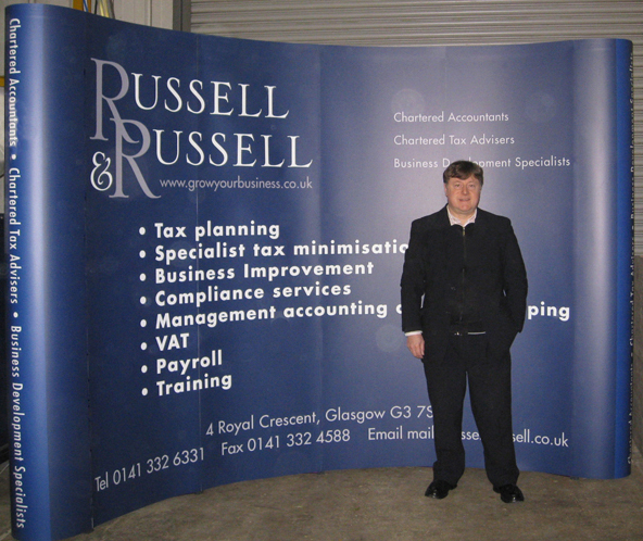 Picture shows a four-by-three size exhibition stand I designed for Russell & Russell Accountants. Wider than most exhibition stands, it's a dark blue curved wall with the R&R logo top left, white bulletpoint text in the centre and contact information along the foot. The curved endcap corners have a lighter shade of blue. I'm shown standing in front of the stand in the workshop.