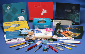 Picture shows a wide range of Promotional Goods supplied by Adamson Design, including branded pens, keyrings, mousemats, pads, USB-sticks and camera. The brightly-coloured gifts are shown on a blue tabletop.