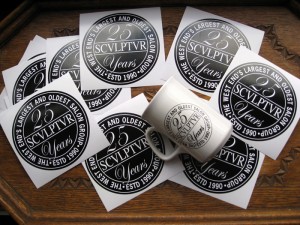 The picture shows a white mug branded with the Sculptur 25 Years Logo on top of adhesive logo-stickers with the same black and white logo. Background is an antique wooden tray.