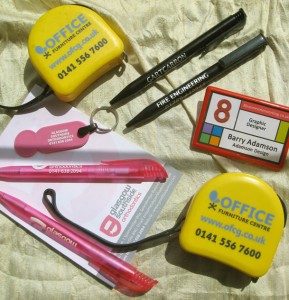Picture shows a variety of Promotional Goods branded with clients' logos and details, including pens, notepads, badges, trolley coin key rings and tape measures.
