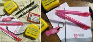 Picture shows a variety of Promotional Goods branded with clients' logos and details, including pens, notepads, badges, trolley coin key rings and tape measures.