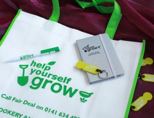 Close-up of promotional goods branded for the Help Yourself Grow project, consisting of tote-bag, pen, notebook with silver cover and trolley-coin key rings.