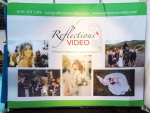 Exhibition stand design: photo of an exhibition stand of the “fabric wall” type, a colourful fabric background stretched over a tubular aluminium frame. This one was designed for Reflections Video (wedding videos).