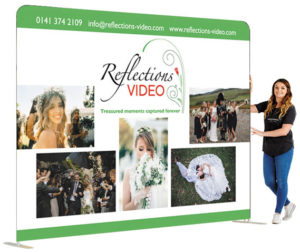 Picture shows a straight wall exhibition stand, 2.3 high x 3 metres wide with a girl standing beside it to show scale. The graphics on it are wedding photos for Reflections Video, wedding videographers.