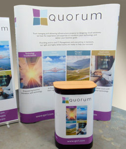 Pic shows a curved wall exhibition stand for Quorum, Edinburgh, with its transit case set up as a counter in front of it. Logo colours are purple, pale blue, teal and ochre with purple on white background for the panels.